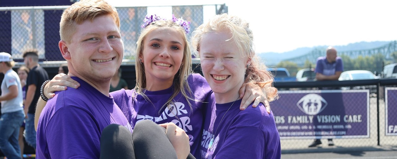 Two students holding a third wearing purple shirts and smiling