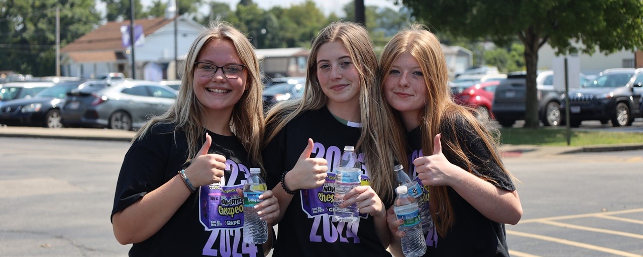 Three girls wearing black and purple shirts with thumbs up smiling
